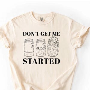 May include: Ivory-colored t-shirt with the text "DON'T GET ME STARTED" and illustrations of three mason jars. The jars have different contents, suggesting a theme of fermentation or food preservation. The shirt is made of a soft material.