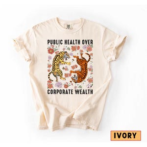 May include: Ivory-colored t-shirt featuring a graphic with two tigers surrounded by flowers. The text "PUBLIC HEALTH OVER CORPORATE WEALTH" is printed above and below the tiger graphic. The shirt is a comfortable, casual style.