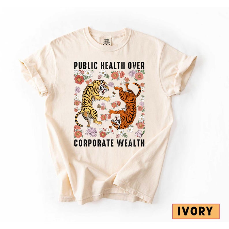 May include: Ivory-colored t-shirt featuring a graphic with two tigers surrounded by flowers. The text "PUBLIC HEALTH OVER CORPORATE WEALTH" is printed above and below the tiger graphic. The shirt is a comfortable, casual style.