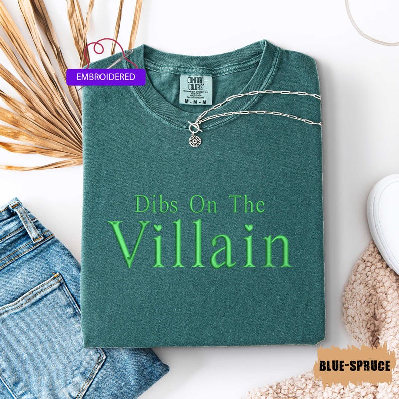 May include: A folded blue-spruce colored t-shirt with the embroidered text "Dibs On The Villain" in green. A silver necklace with a circular pendant rests on the shirt. The word "EMBROIDERED" is in a purple rectangle.