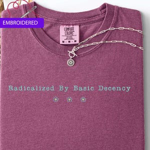 May include: A maroon t-shirt with the text "Radicalized By Basic Decency" in light blue. Below the text are three small embroidered flowers. A silver necklace with a floral pendant rests on the shirt. The tag reads "Comfort Colors".
