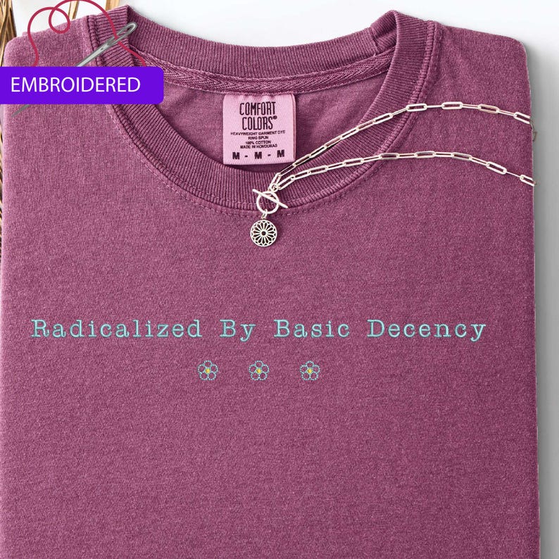 May include: A maroon t-shirt with the text "Radicalized By Basic Decency" in light blue. Below the text are three small embroidered flowers. A silver necklace with a floral pendant rests on the shirt. The tag reads "Comfort Colors".