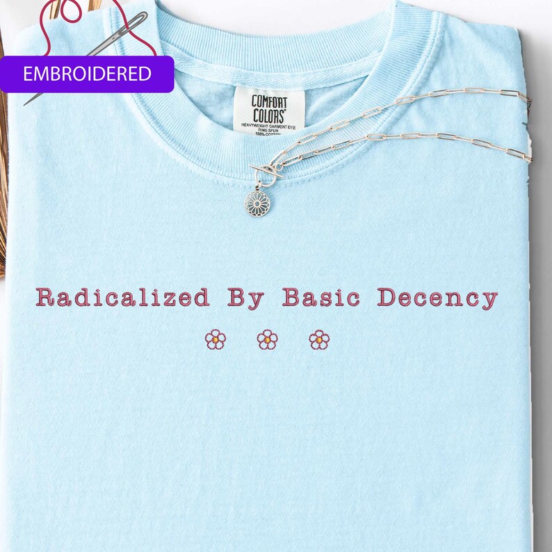 May include: Light blue t-shirt with the embroidered text "Radicalized By Basic Decency" in a reddish-brown color. Below the text are three small embroidered flowers. A silver necklace with a flower pendant rests on the shirt.