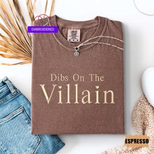 May include: Espresso-colored t-shirt with the phrase "Dibs On The Villain" in a serif font. The shirt has a crew neck and short sleeves. A silver necklace with a sunburst pendant is on the shirt. The word "Embroidered" is on a purple tag.