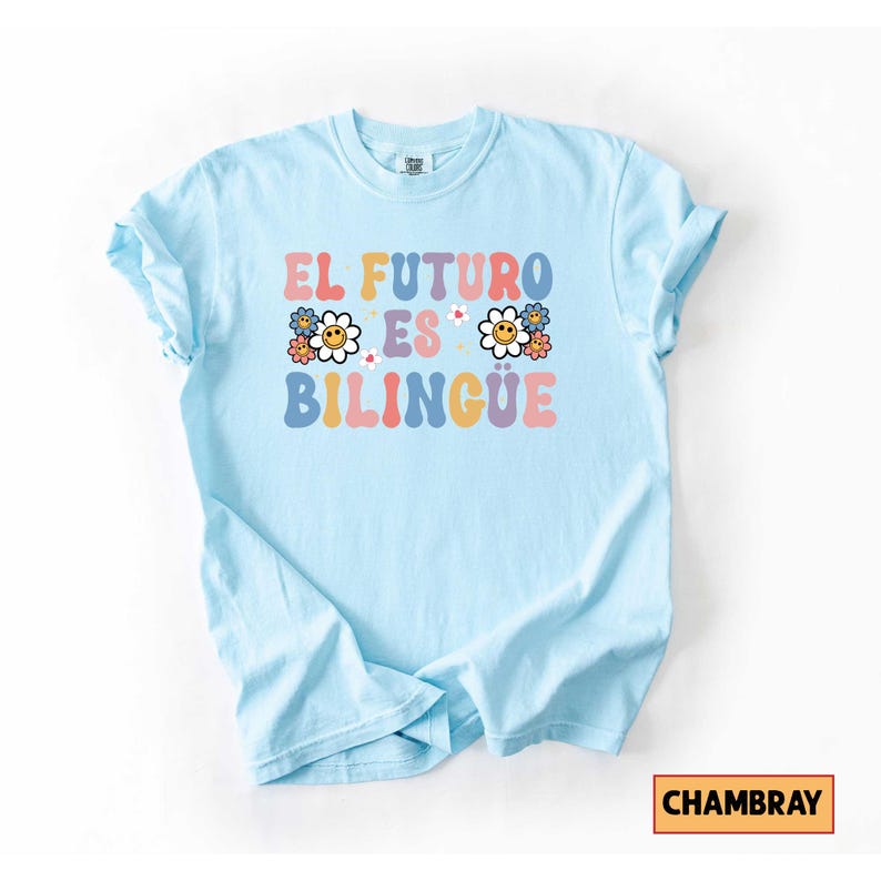Pu&ograve; includere: Una maglietta azzurra con la scritta "EL FUTURO ES BILINGUE" in lettere colorate in stile retr&ograve;. Il design include grafiche floreali. La maglietta &egrave; realizzata in materiale morbido ed &egrave; piegata su una superficie bianca.