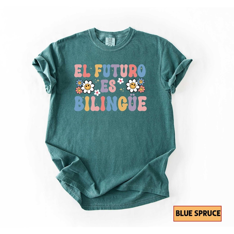 Pu&ograve; includere: Una maglietta color verde acqua con la frase "EL FUTURO ES BILINGUE" in lettere colorate in stile retr&ograve;. Il design include margherite e grafiche floreali. La maglietta &egrave; etichettata "Blue Spruce".