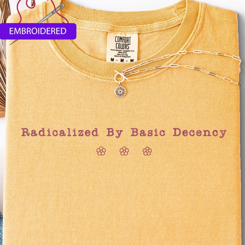 May include: Mustard yellow embroidered t-shirt with the text "Radicalized By Basic Decency" and three flower graphics. A silver necklace with a floral pendant rests on the shirt. The shirt is labeled "Comfort Colors".