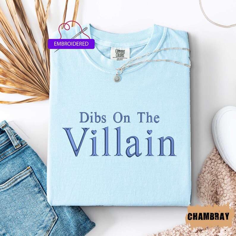 May include: A light blue t-shirt with the embroidered text "Dibs On The Villain" in a darker blue font. The shirt is folded and displayed with a pair of blue jeans and a silver necklace. The shirt has the "Comfort Colors" brand label.
