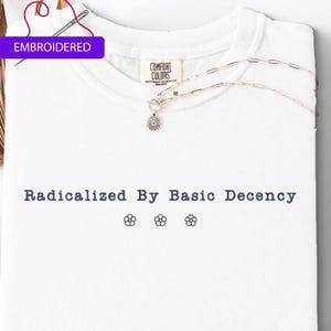 May include: White t-shirt with the phrase "Radicalized By Basic Decency" in navy blue, along with three small flower designs. The shirt has a crew neck and a tag that says "Comfort Colors". A silver necklace with a flower pendant is on the shirt.