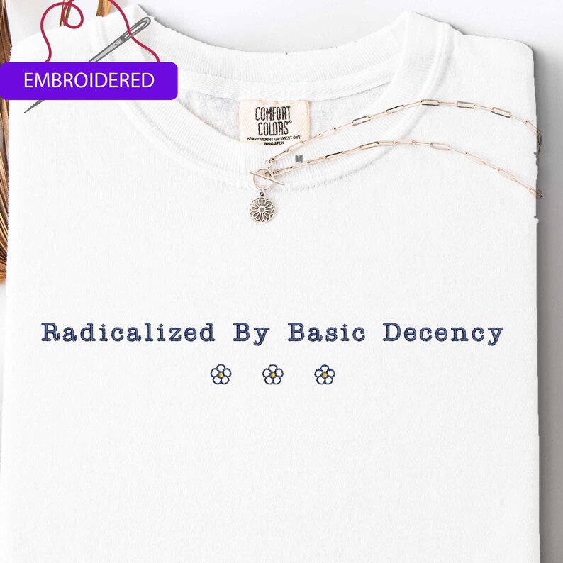May include: White t-shirt with the phrase "Radicalized By Basic Decency" in navy blue, along with three small flower designs. The shirt has a crew neck and a tag that says "Comfort Colors". A silver necklace with a flower pendant is on the shirt.