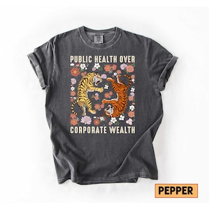 May include: Dark gray t-shirt with the text "PUBLIC HEALTH OVER CORPORATE WEALTH." The shirt features two tigers surrounded by flowers. One tiger is upright, and the other is upside down. The shirt is a crew neck.