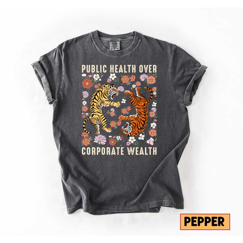 May include: Dark gray t-shirt with the text "PUBLIC HEALTH OVER CORPORATE WEALTH." The shirt features two tigers surrounded by flowers. One tiger is upright, and the other is upside down. The shirt is a crew neck.