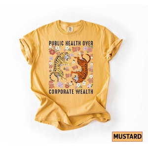 May include: Mustard-colored t-shirt with the text "PUBLIC HEALTH OVER CORPORATE WEALTH." The graphic features two tigers surrounded by flowers. The shirt is a solid color with a crew neck and short sleeves.