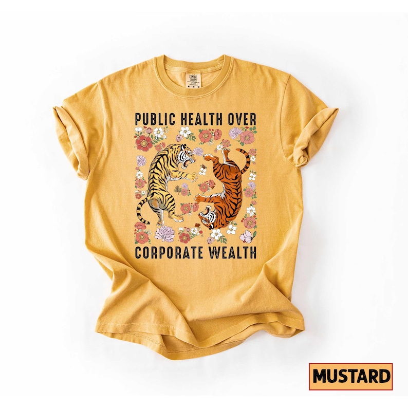 May include: Mustard-colored t-shirt with the text "PUBLIC HEALTH OVER CORPORATE WEALTH." The graphic features two tigers surrounded by flowers. The shirt is a solid color with a crew neck and short sleeves.