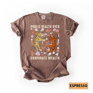 May include: Espresso-colored t-shirt with the text "PUBLIC HEALTH OVER CORPORATE WEALTH." The shirt features a graphic of two tigers surrounded by flowers. One tiger is yellow and black, the other is orange and black.