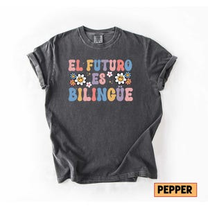 Pu&ograve; includere: T-shirt grigio scuro con la scritta "EL FUTURO ES BILINGUE" in lettere colorate in stile retr&ograve;. Il design include fiori e faccine sorridenti, creando un tema allegro ed educativo. La maglietta &egrave; realizzata in materiale morbido.