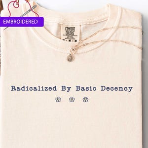 May include: Cream-colored t-shirt with the phrase "Radicalized By Basic Decency" in navy blue, along with three embroidered flower designs. The shirt has a Comfort Colors tag and a silver necklace. The word "EMBROIDERED" is in a purple box.