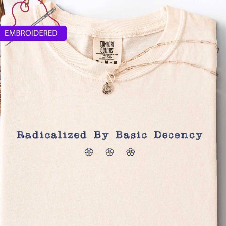 May include: Cream-colored t-shirt with the phrase "Radicalized By Basic Decency" in navy blue, along with three embroidered flower designs. The shirt has a Comfort Colors tag and a silver necklace. The word "EMBROIDERED" is in a purple box.