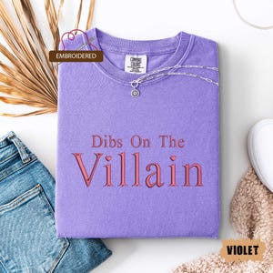 May include: A violet-colored t-shirt with the embroidered text "Dibs On The Villain" in a pink hue. The shirt is folded, with a silver necklace and a small pendant visible. The word "Embroidered" is in a brown box.