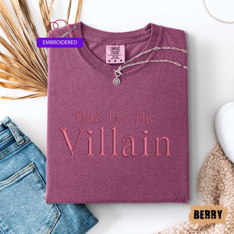 May include: A berry-colored t-shirt with the embroidered text "Dibs On The Villain". The shirt is folded and has a silver necklace with a pendant. A tag that says "Embroidered" is visible.