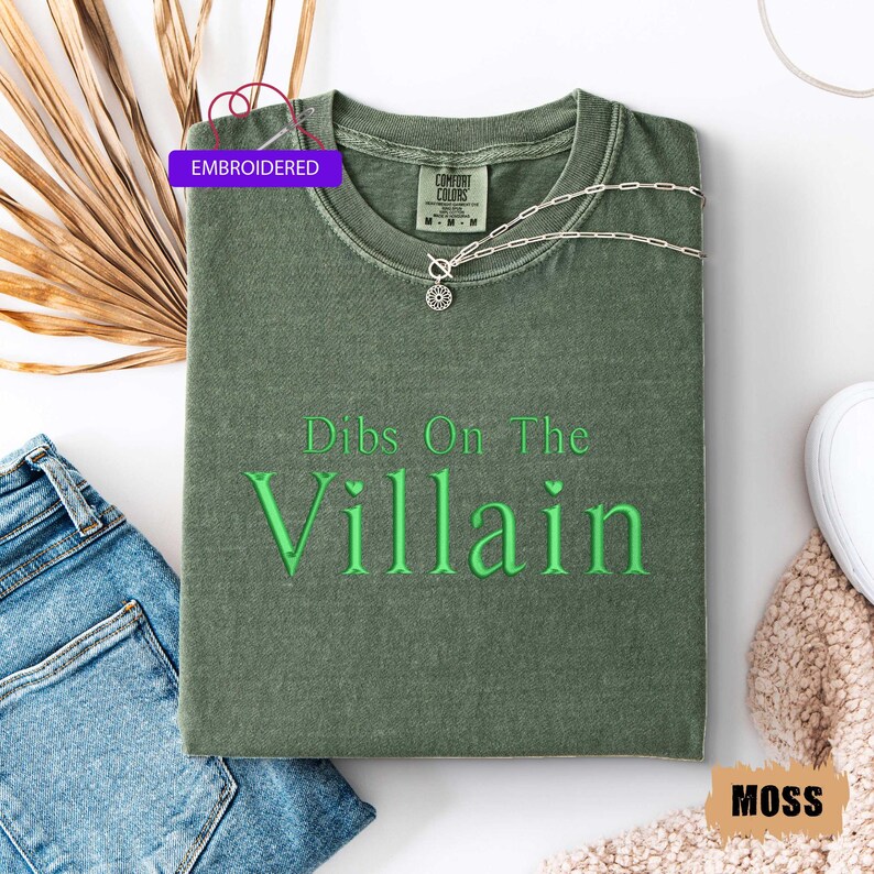 May include: A moss green t-shirt with the embroidered text "Dibs On The Villain" in green. The shirt is folded and has a silver necklace with a pendant. The shirt is next to a pair of blue jeans and a white shoe.
