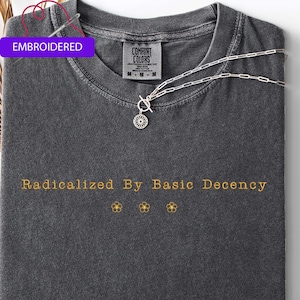 May include: Dark gray t-shirt with the embroidered text "Radicalized By Basic Decency" in gold. The shirt has a crew neck and a Comfort Colors tag. A silver necklace with a floral pendant is draped across the shirt.