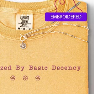 May include: Mustard yellow t-shirt with the embroidered text "zed By Basic Decency" and three flower designs. A silver necklace with a floral pendant and a toggle clasp is draped across the shirt. The word "EMBROIDERED" is in a purple rectangle.