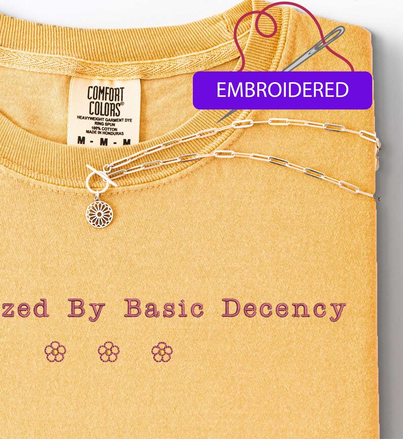 May include: Mustard yellow t-shirt with the embroidered text "zed By Basic Decency" and three flower designs. A silver necklace with a floral pendant and a toggle clasp is draped across the shirt. The word "EMBROIDERED" is in a purple rectangle.