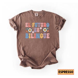 Pu&ograve; includere: T-shirt color espresso con la scritta "EL FUTURO ES BILING&Uuml;E" in stile retr&ograve; colorato, con dettagli floreali. La maglietta &egrave; di stile casual e confortevole.
