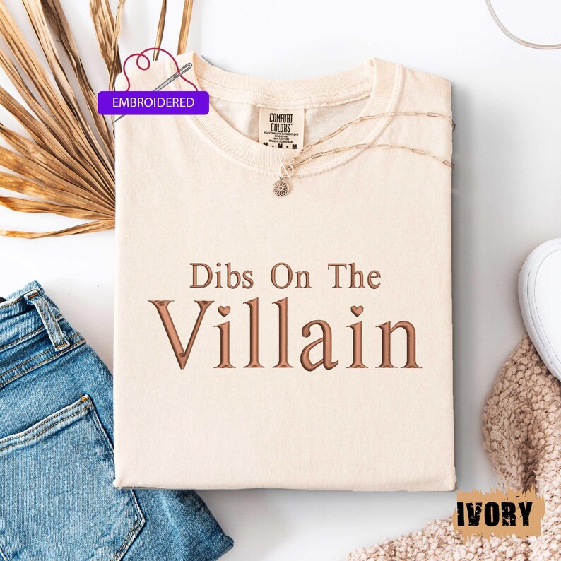 May include: Ivory-colored t-shirt with the embroidered text "Dibs On The Villain" in brown. The shirt is folded and has a Comfort Colors tag. A gold necklace and a pair of blue jeans are next to the shirt.
