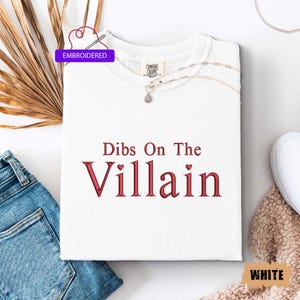 May include: White t-shirt with the embroidered text "Dibs On The Villain" in red. The shirt has a crew neck and is folded neatly. A silver necklace with a pendant rests on the shirt. The word "Embroidered" is in a purple box.