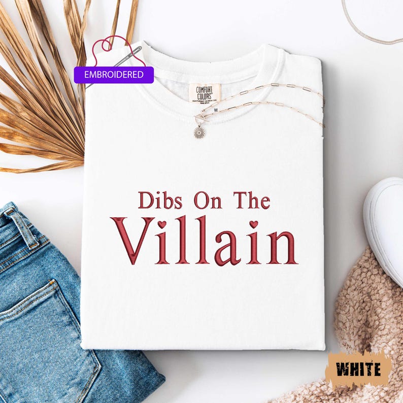 May include: White t-shirt with the embroidered text "Dibs On The Villain" in red. The shirt has a crew neck and is folded neatly. A silver necklace with a pendant rests on the shirt. The word "Embroidered" is in a purple box.