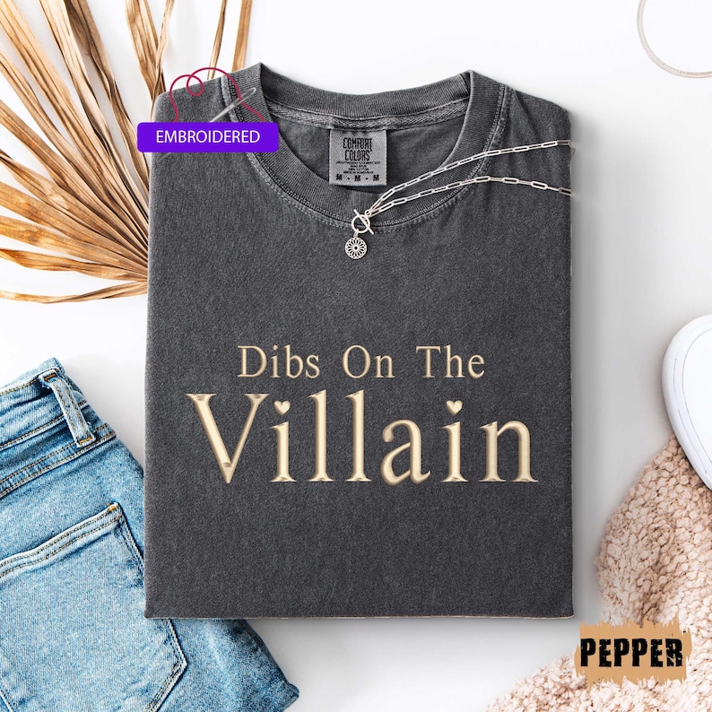May include: A dark gray t-shirt with the phrase "Dibs On The Villain" in a gold-colored font. The shirt is styled with a silver necklace and a pair of blue jeans. The word "Embroidered" is on a purple tag.