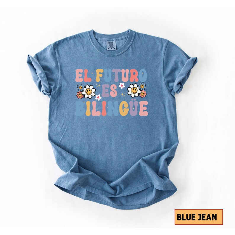 Pu&ograve; includere: Una maglietta blu jeans con la scritta "EL FUTURO ES BILINGUE" in lettere colorate in stile retr&ograve;. Il design include grafiche floreali. La maglietta &egrave; piegata, con le maniche arrotolate.