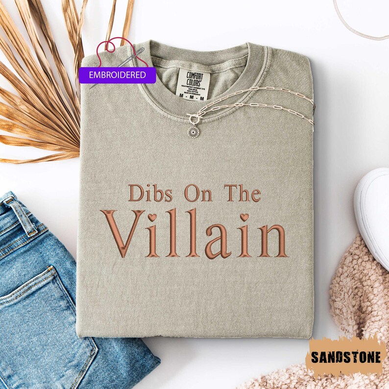 May include: Sandstone-colored t-shirt with the embroidered phrase "Dibs On The Villain" in a copper-colored font. The shirt is folded and accessorized with a silver necklace. The tag reads "Embroidered".