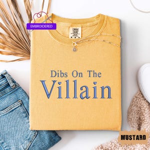 May include: Mustard-colored t-shirt with the embroidered text "Dibs On The Villain" in blue. The shirt has a crew neck and is folded. A gold necklace with a sun pendant is on top of the shirt. The word "Embroidered" is in a purple banner.
