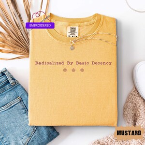 May include: Mustard-colored embroidered t-shirt with the text "Radicalized By Basic Decency" and three small flower graphics. A silver necklace with a sun pendant is draped across the shirt. The shirt is folded on a white surface next to a pair of blue jeans.