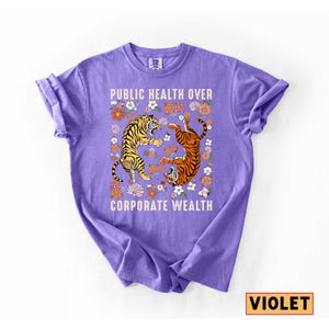 May include: Violet-colored t-shirt with the text "PUBLIC HEALTH OVER CORPORATE WEALTH." The shirt features two tigers surrounded by floral designs. One tiger is yellow and black, the other is orange and black.