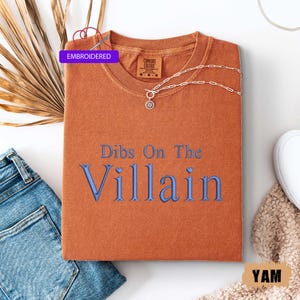 May include: An orange Comfort Colors t-shirt with the embroidered text "Dibs On The Villain" in blue. A silver necklace with a pendant is draped on the shirt. The word "Embroidered" is in a purple box.
