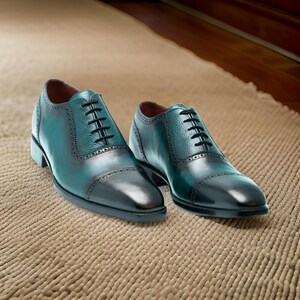 May include: A pair of dark green leather lace-up shoes with a cap toe design. The shoes are on a light brown textured surface.