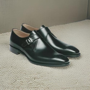 Könnte beinhalten: Ein Paar schwarze Monk-Strap-Schuhe aus Leder mit Brogue-Muster. Die Schuhe haben einen Schnallenverschluss und eine spitze Zehenpartie.