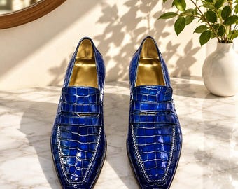 May include: A pair of vibrant blue loafers featuring a glossy, embossed crocodile skin texture. These shoes have a classic penny loafer strap across the vamp, a pointed toe silhouette, and a smooth, metallic gold-toned interior lining.