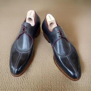 Könnte beinhalten: Ein Paar dunkelblaue Lederschuhe mit spitzer Zehenpartie und Schnürung. Die Schuhe haben einen kontrastierenden hellbraunen Lederbesatz an den Rändern und eine dunkelbraune Ledersohle.