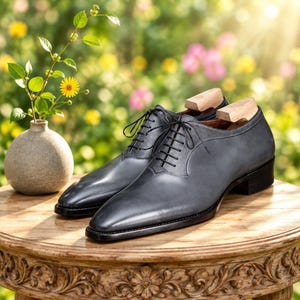 May include: A pair of dark gray leather oxford shoes with black laces and wooden shoe trees inserted. The shoes feature a smooth toe cap and a textured leather quarter panel, resting on a wooden table next to a small ceramic vase with flowers.