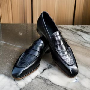 May include: A pair of black leather loafers with a crocodile-embossed design. The loafers have a square toe and a slip-on style.