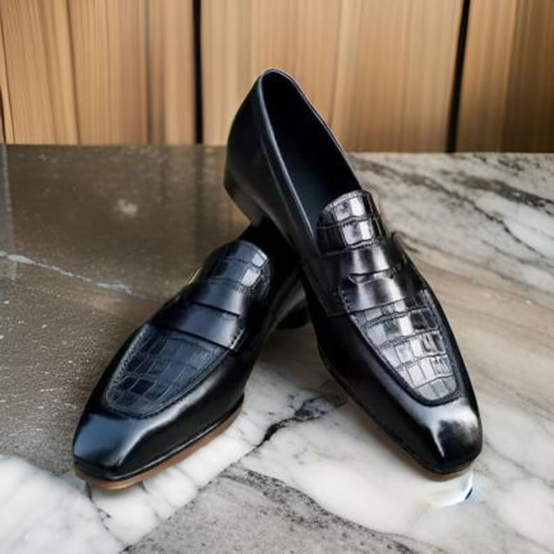 May include: A pair of black leather loafers with a crocodile-embossed design. The loafers have a square toe and a slip-on style.