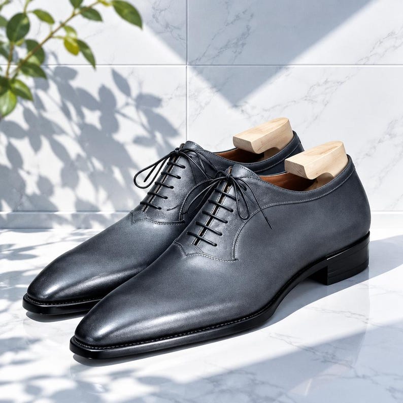 May include: A pair of dark gray leather oxford shoes with black laces, a pointed toe, and a low black heel. Each shoe contains a light-colored wooden shoe tree insert to maintain the shape of the footwear while displayed on a white surface.