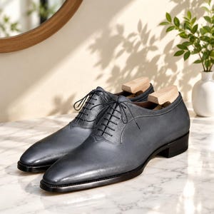 May include: A pair of dark grey leather oxford shoes featuring a smooth toe cap, textured leather lace panels, thin black laces, and a low black heel. Each shoe contains a light-colored wooden shoe tree inserted to maintain the shape of the footwear.