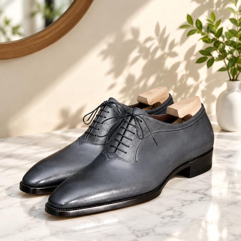 May include: A pair of dark grey leather oxford shoes featuring a smooth toe cap, textured leather lace panels, thin black laces, and a low black heel. Each shoe contains a light-colored wooden shoe tree inserted to maintain the shape of the footwear.