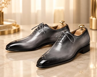 May include: A pair of oxford shoes featuring a polished grey and black gradient finish, a pointed toe, and matching grey laces. Each shoe includes a light-colored wooden shoe tree with a gold-toned metal knob inserted into the heel area.